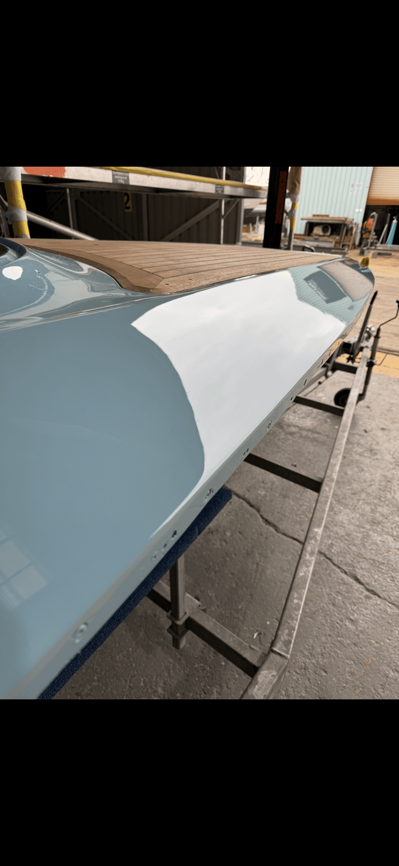 Reflective light blue boat hull with a wooden deck, resting on a stand in a workshop.