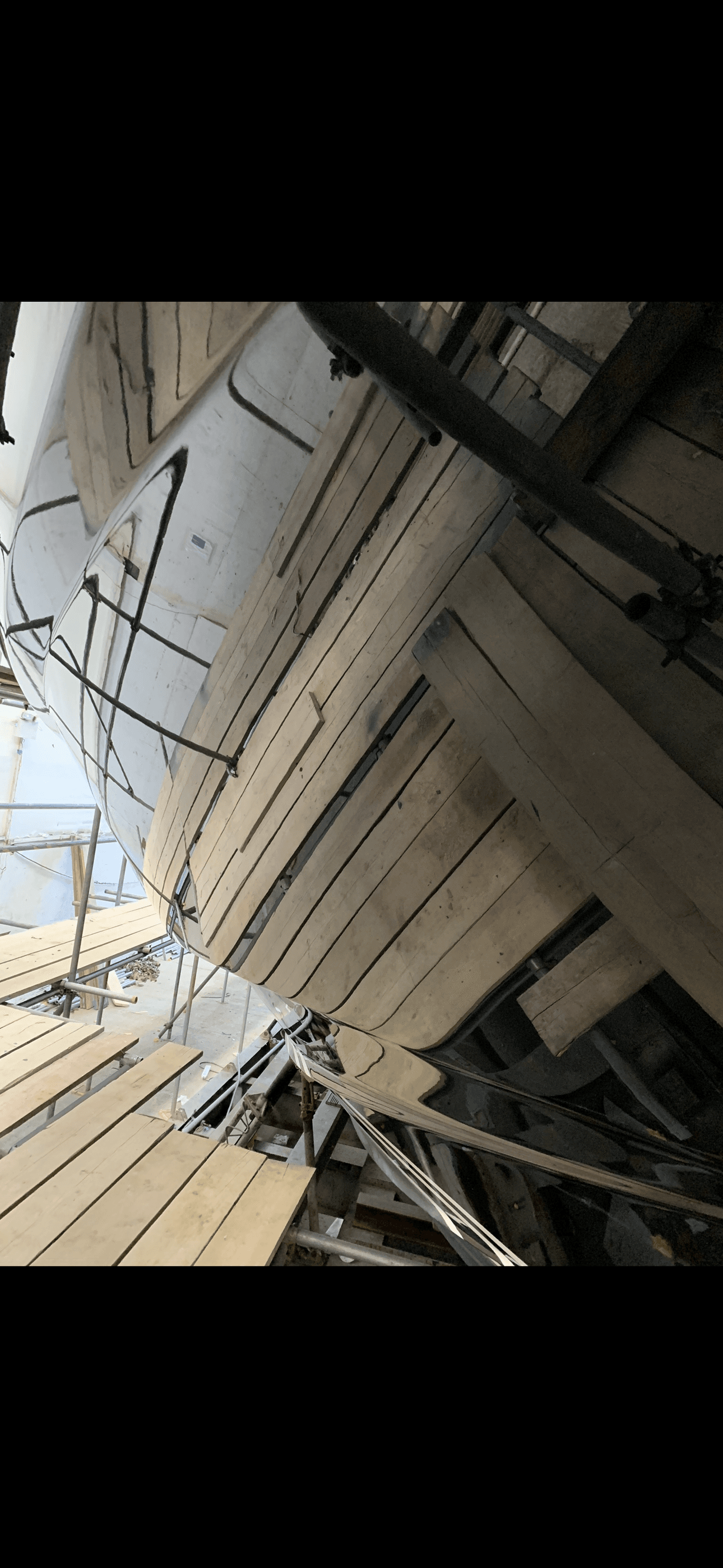 Smooth, reflective boat hull under construction, surrounded by wooden planks and metal scaffolding.